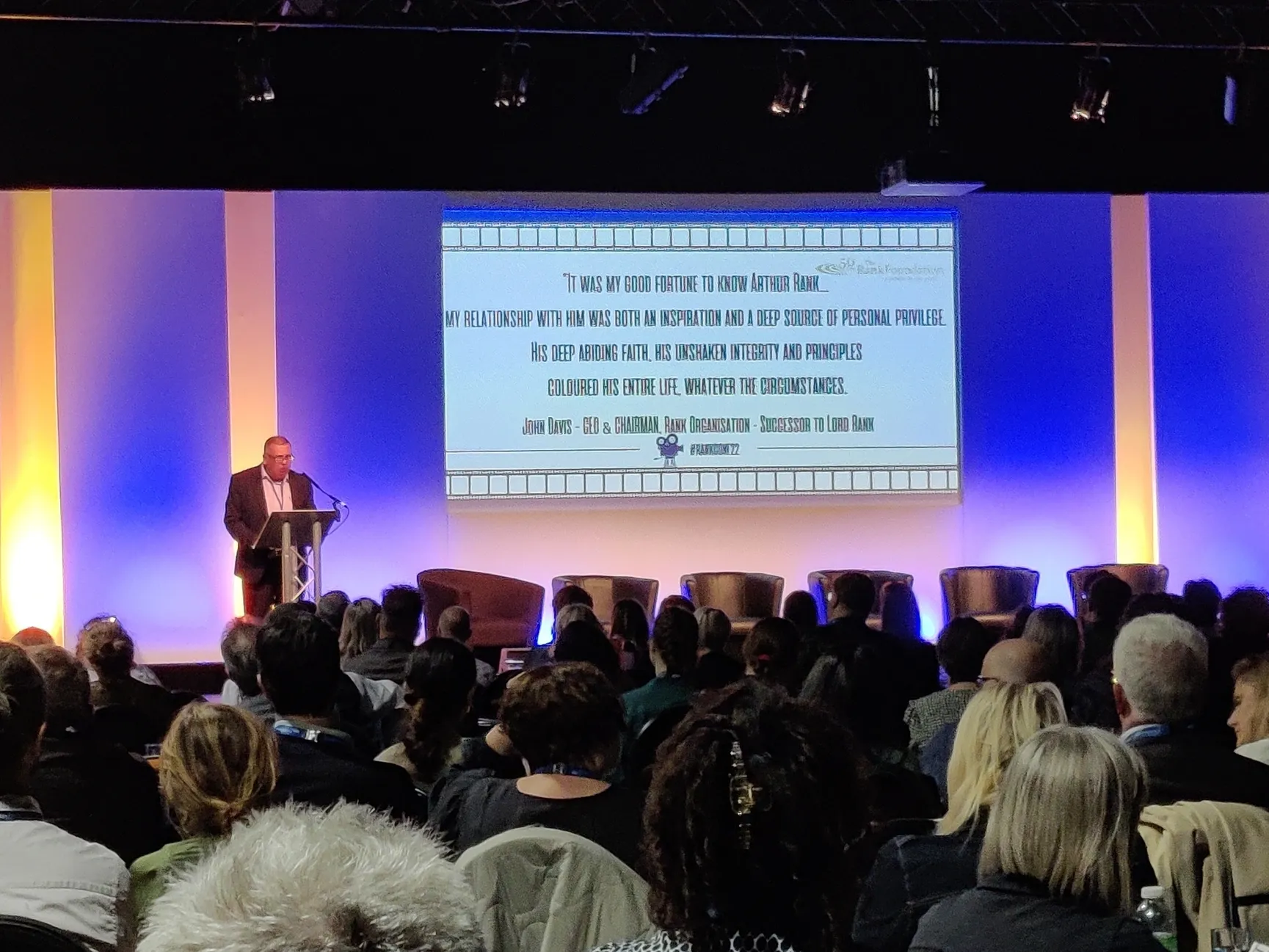 Conference production setup with stage lighting, projection, and a speaker at a lectern.